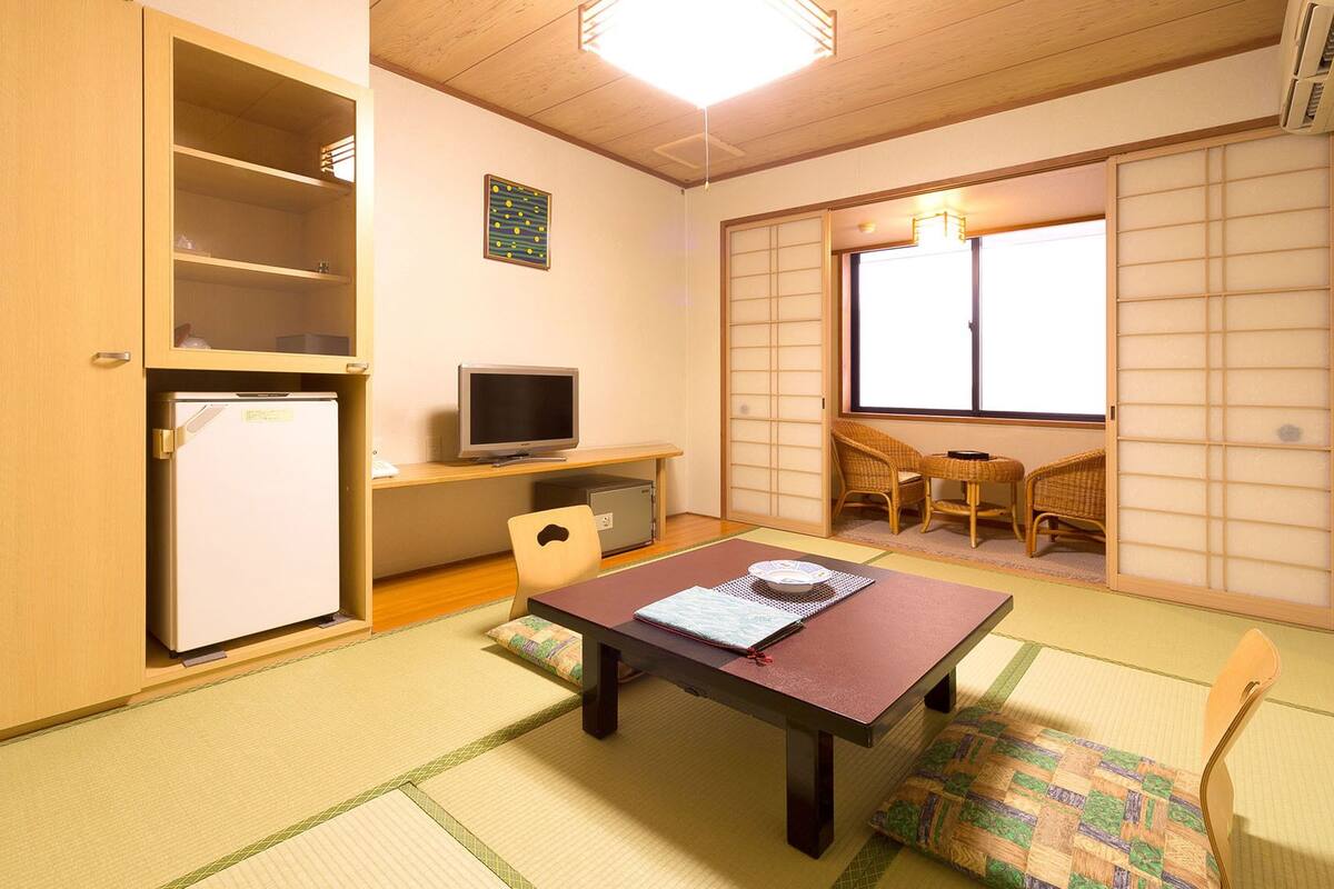 Ein Zimmer mit einem Holztisch, einem Fernseher, einem kleinen Kühlschrank, einem Tatami-Boden und einer schiebbaren Shoji-Schiebetür.