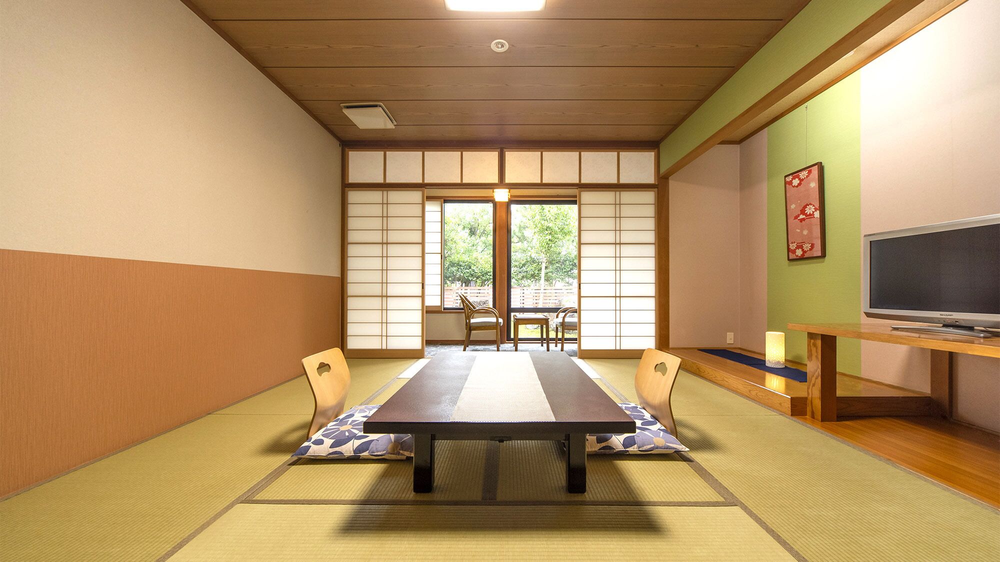 Habitació japonesa tradicional amb sòcols de tatami, una taula baixa, cadires, un televisor i una porta corredissa que dona a una zona exterior.
