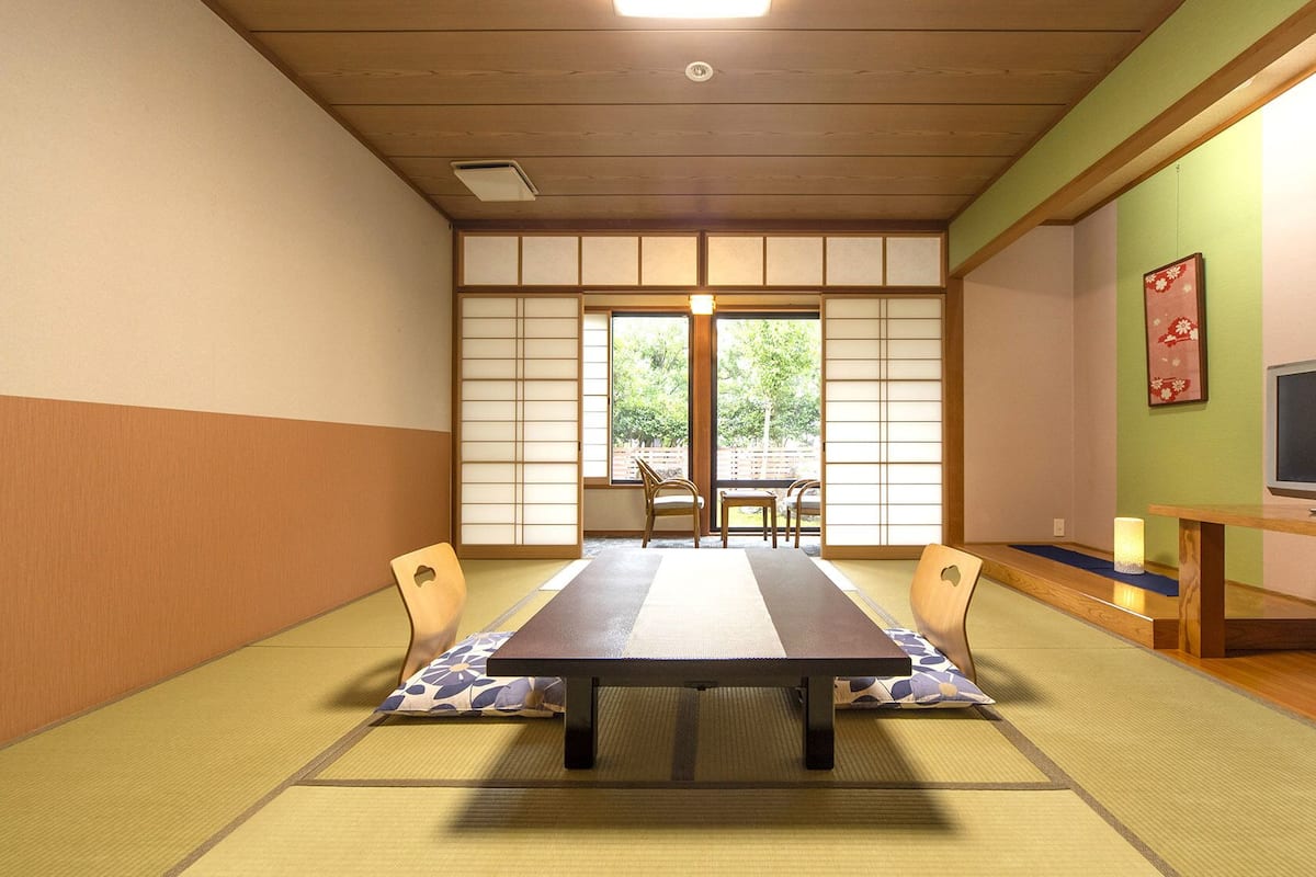 Ein traditioneller japanischer Raum mit Tatami-Boden, einem niedrigen Tisch, Stühlen, einem Fernseher und einer Schiebetür zu einem Außenbereich.