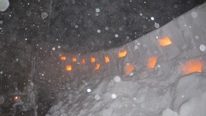 Paisaje nevado con una carretera parcialmente visible y luces anaranjadas.