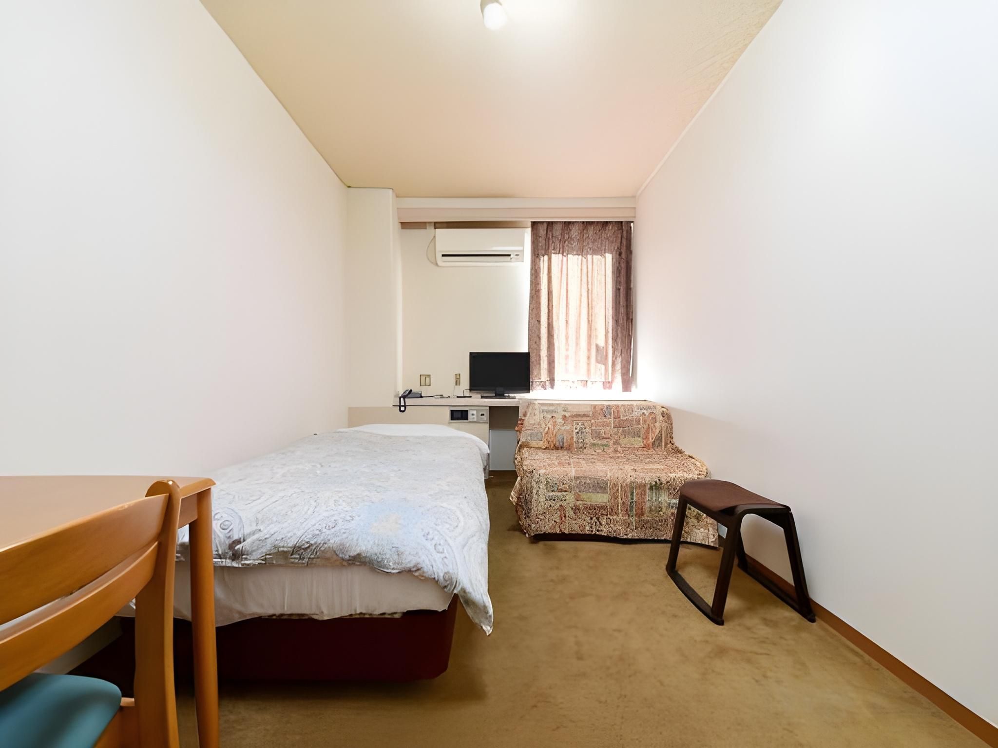 A small, well-lit room with a single bed, a desk, a chair, and a television.