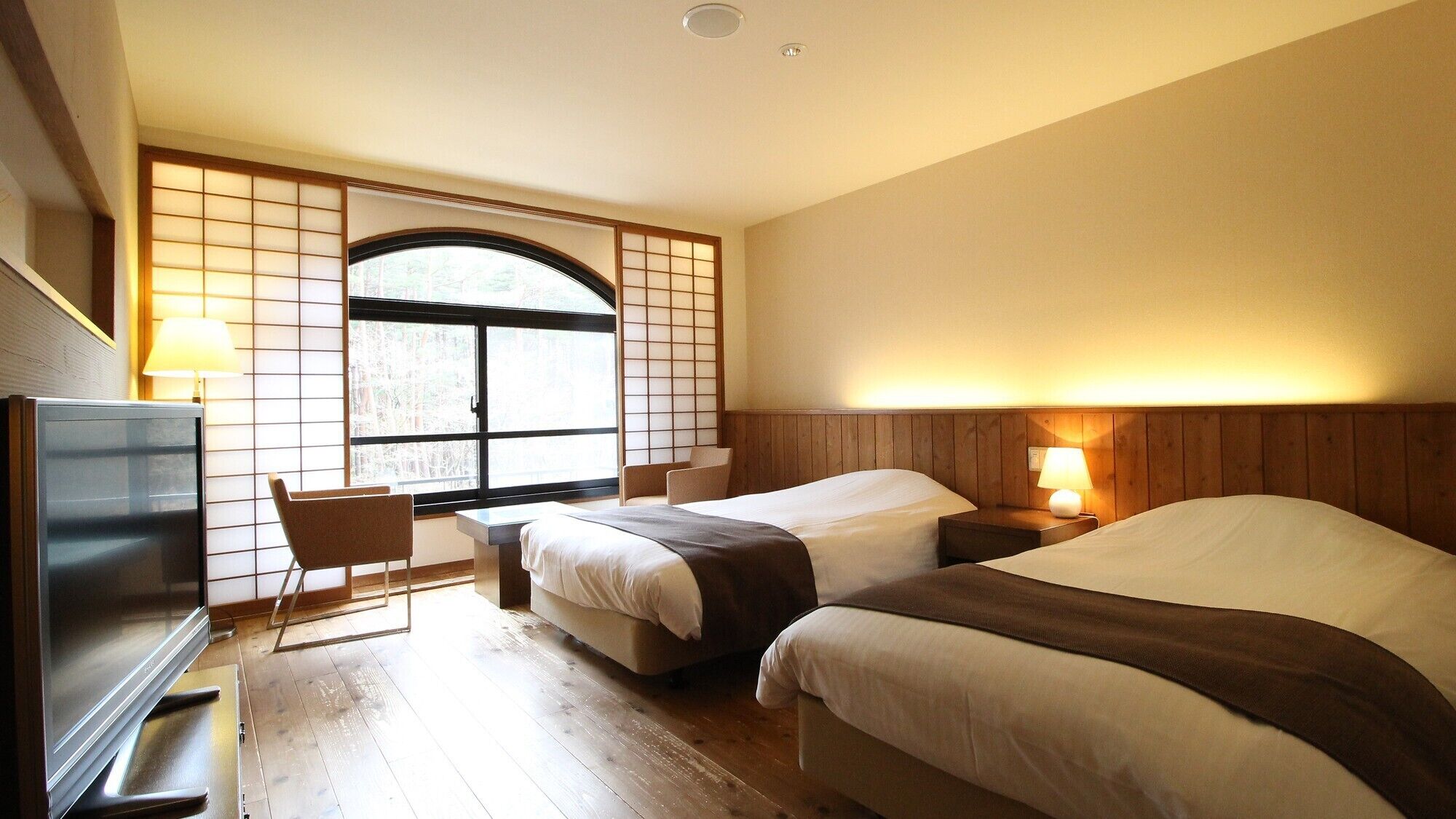 A hotel room with two beds, a desk, a TV, and a window with a view of trees.