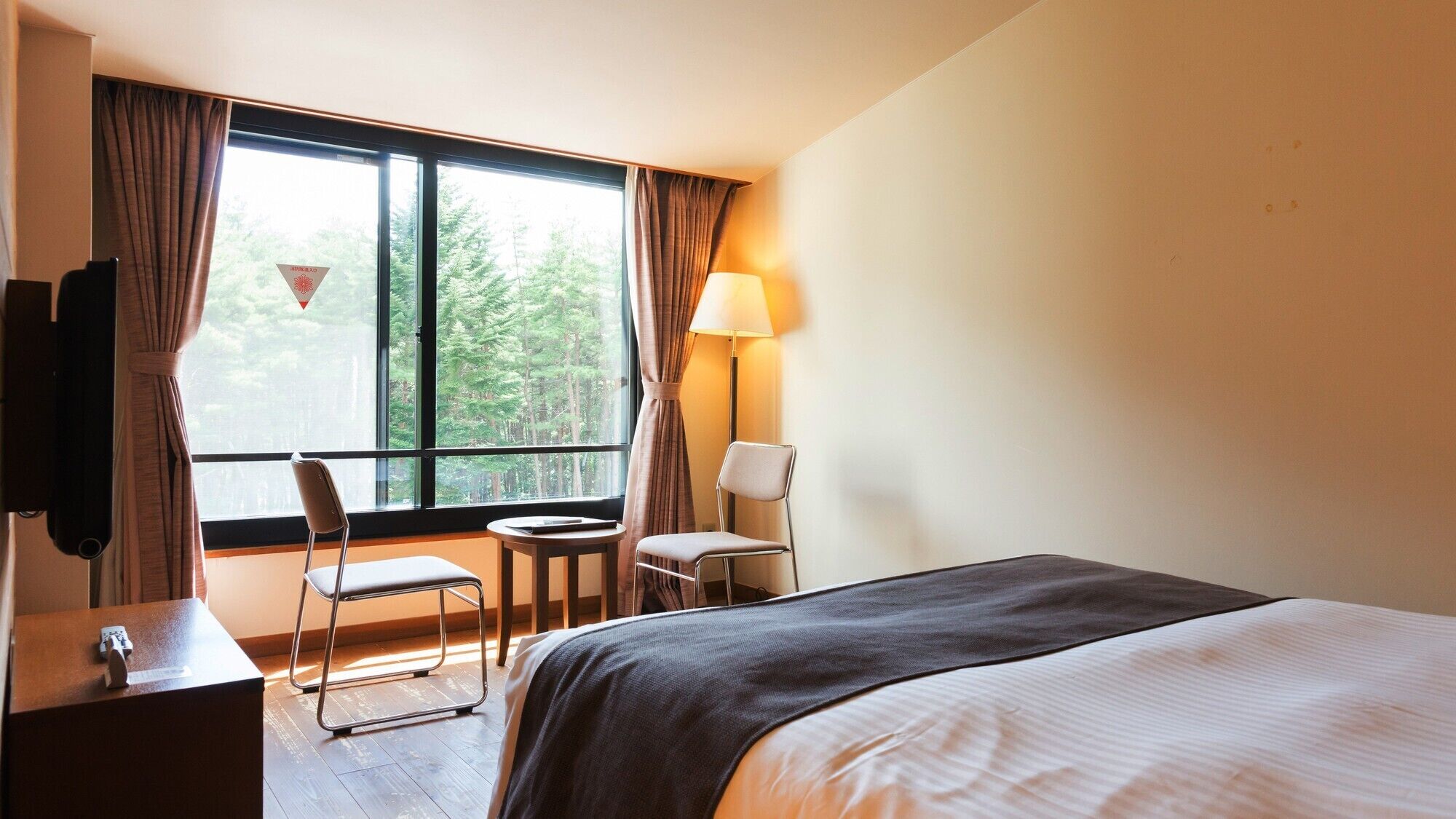 A hotel room with a bed, a desk, a chair, a lamp, and a window with curtains.