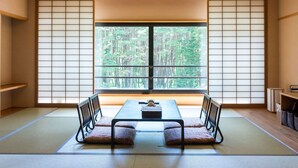 A tatami mat room with a low table and two chairs, a large window with a view of a forest, and a traditional sliding door. - Hotel Fuuka (Hokuto)