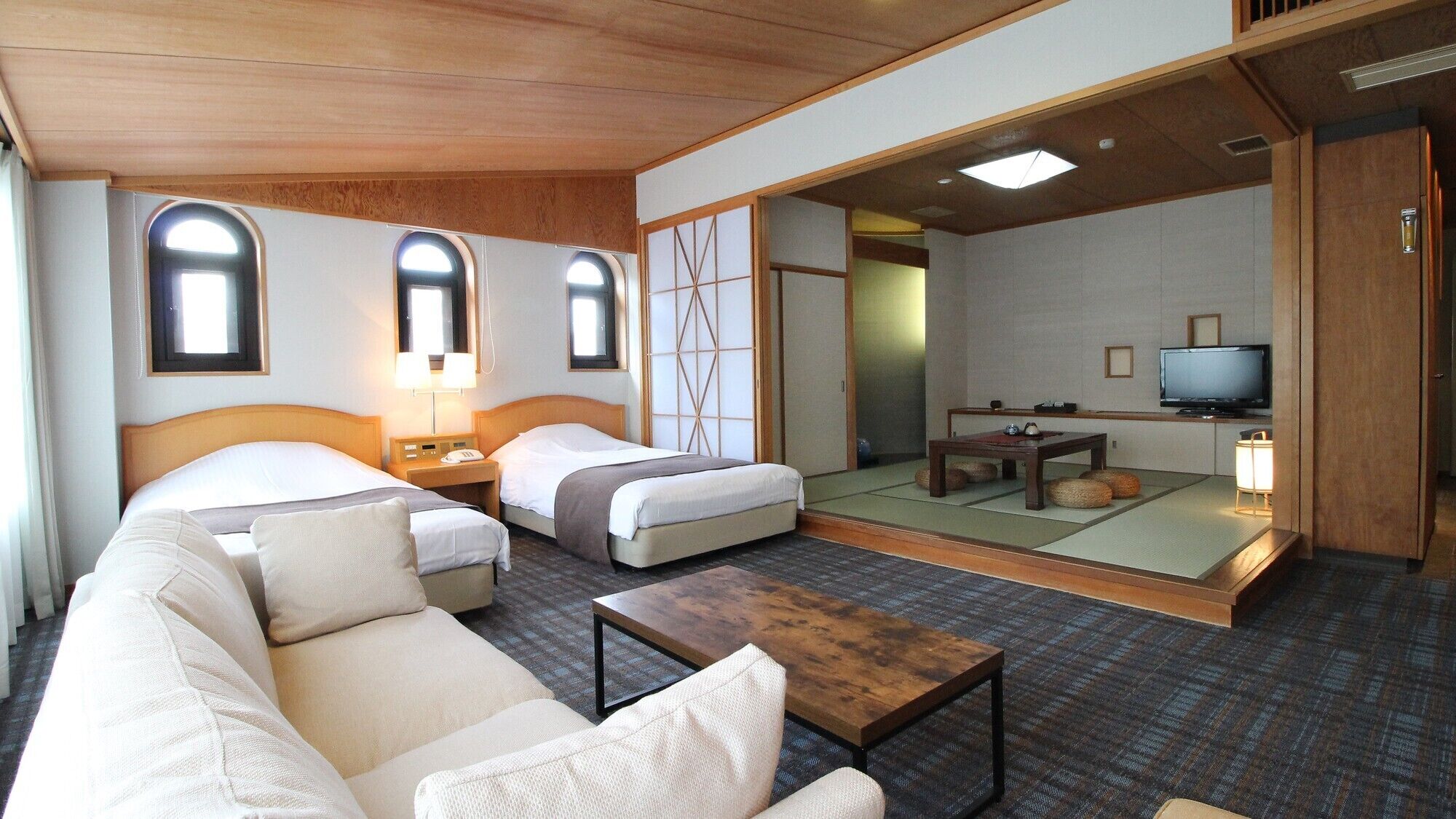 Ein Hotelzimmer mit zwei Betten, einem Sofa, einem kleinen Tisch, einem Fernseher und einer traditionellen japanischen Schiebetür.