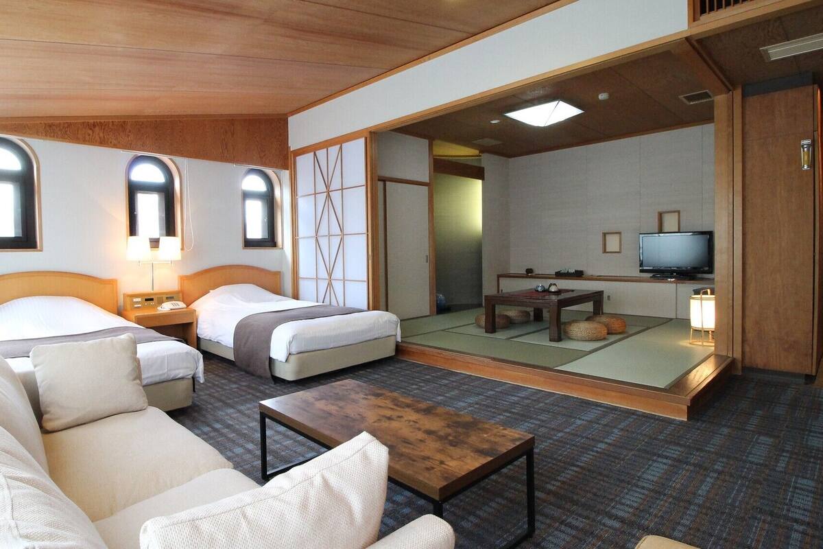 Ein Hotelzimmer mit zwei Betten, einem Sofa, einem kleinen Tisch, einem Fernseher und einer traditionellen japanischen Schiebetür.