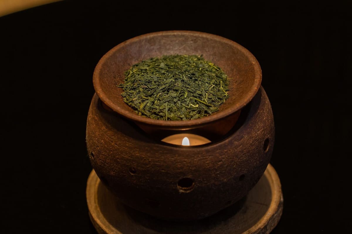A small, round, dark brown ceramic bowl filled with green leaves, placed on a wooden base with a lit candle inside.