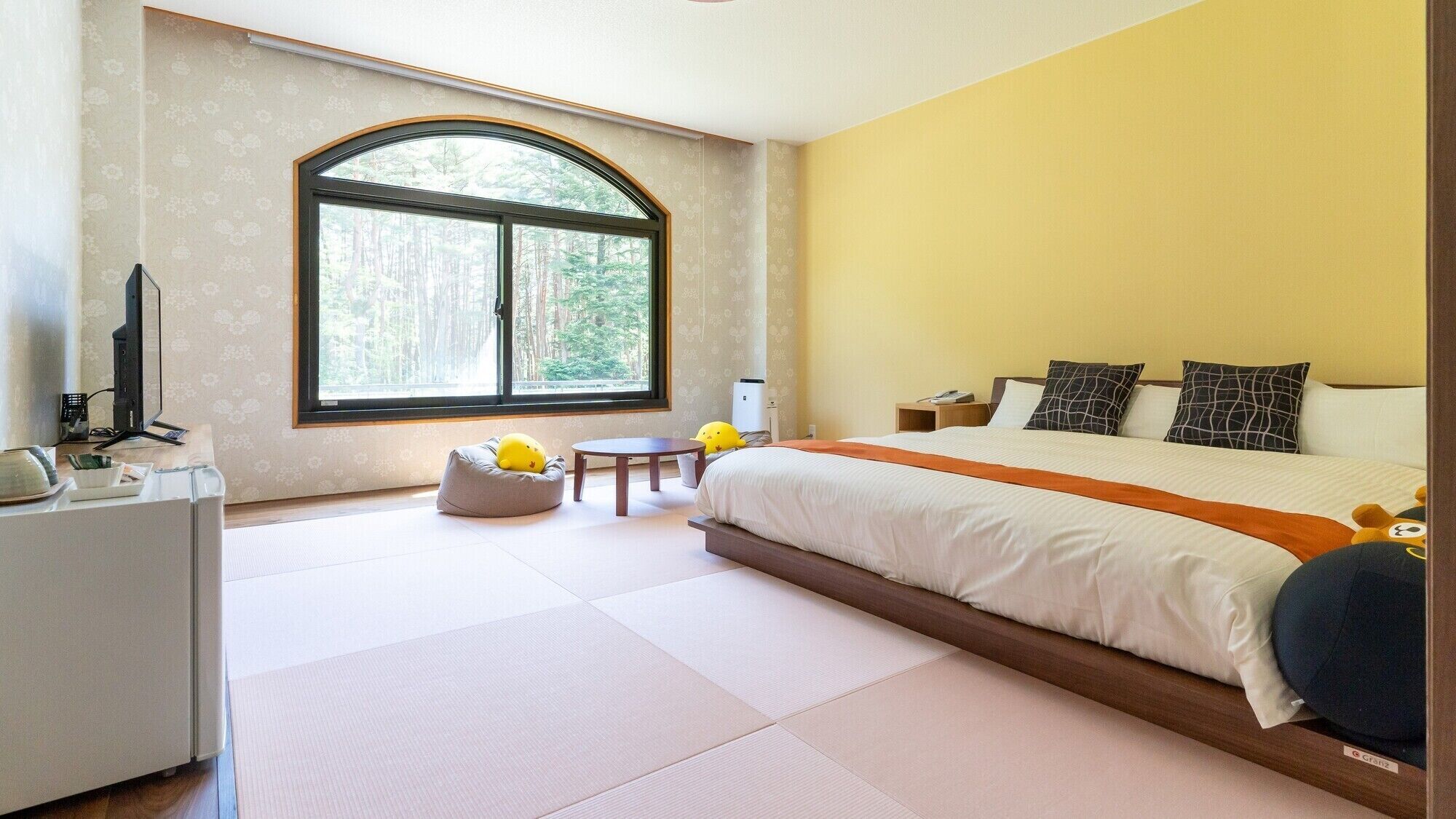 A bedroom with a large bed, a desk with a computer, a chair, and a window with a view of trees.