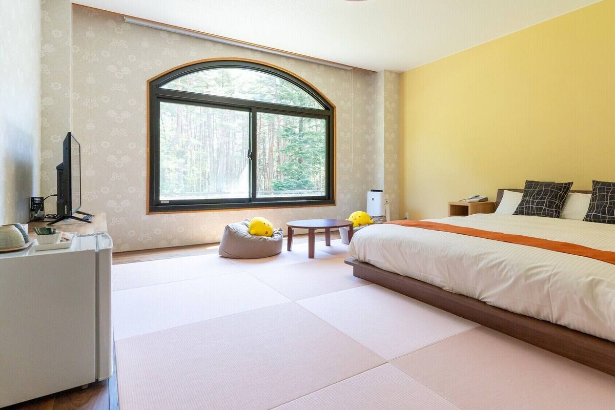 Ein Schlafzimmer mit einem großen Bett, einem Schreibtisch mit Computer, einem Stuhl und einem Fenster mit Blick auf Bäume.