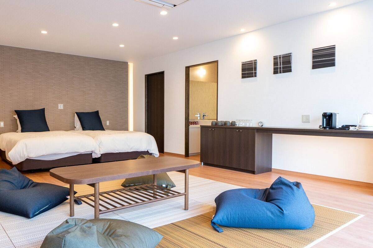 Ein modernes Hotelzimmer mit zwei Betten, einer kleinen Küchenzeile, einem Esstisch und einer Sitzecke mit Sitzsäcken.