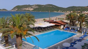 A swimming pool with lounge chairs and umbrellas, surrounded by palm trees and a view of the sea and mountains. - Alkyon Beach Hotel (Corfu)