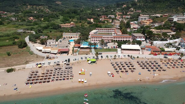 An aerial view of a coastal town with a beach, clear waters, and a mix of residential and commercial buildings. - Alkyon Beach Hotel (Corfu)