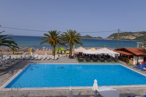 A swimming pool with lounge chairs and umbrellas, a beach in the background, and a bar area. - Alkyon Beach Hotel (Corfu)