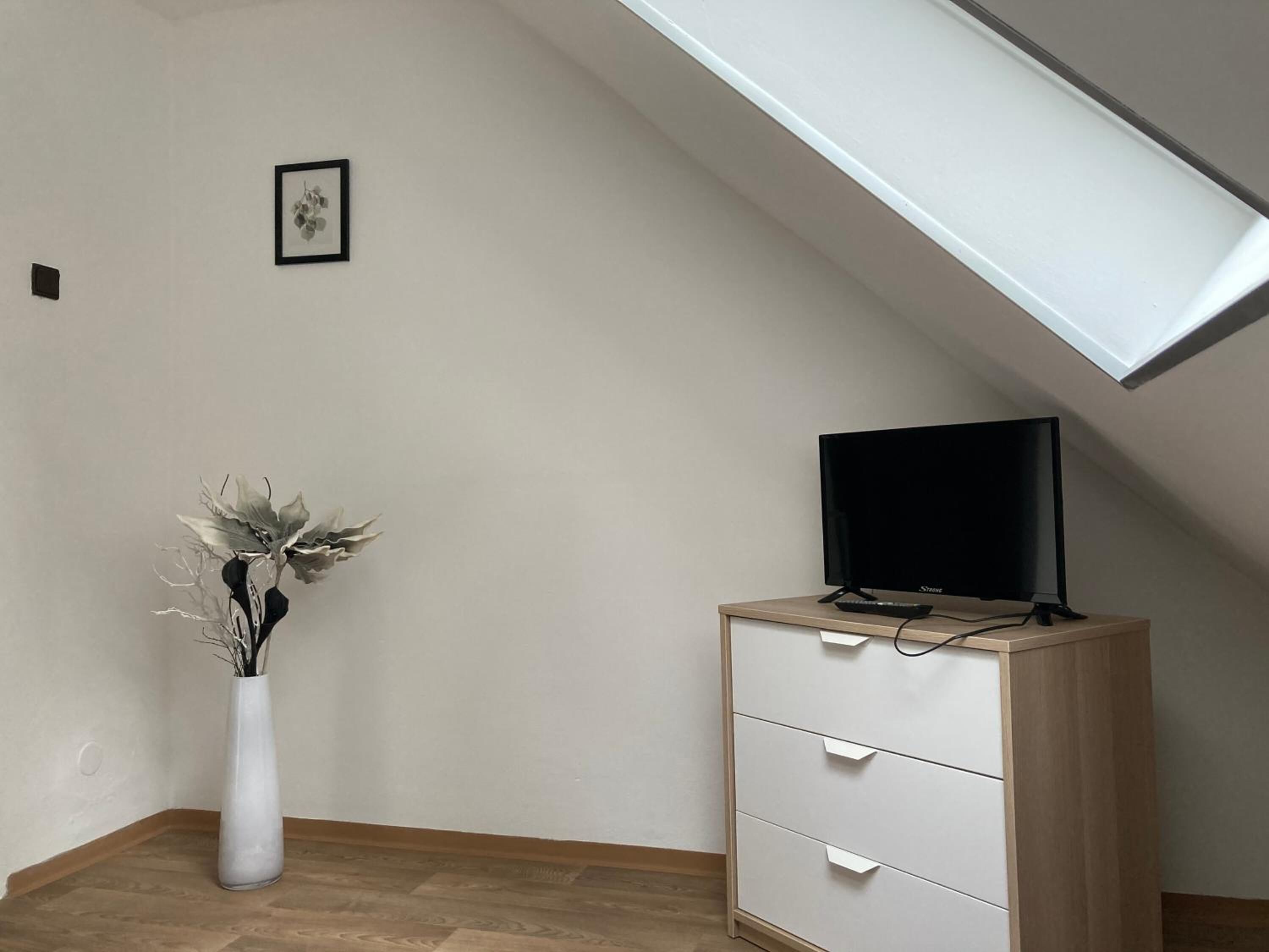 Una habitación con techo inclinado, un escritorio blanco con cajones, un monitor negro y un jarrón blanco con flores secas.