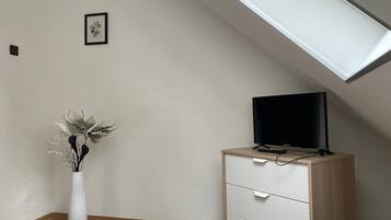 Una habitación con techo inclinado, un escritorio blanco con cajones, un monitor negro y un jarrón blanco con flores secas.