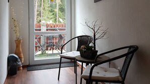 Una habitación con piso de madera, una puerta que da a un espacio exterior con vista a árboles y una cerca, una silla con cojín, una planta en maceta y un cuadro en la pared.