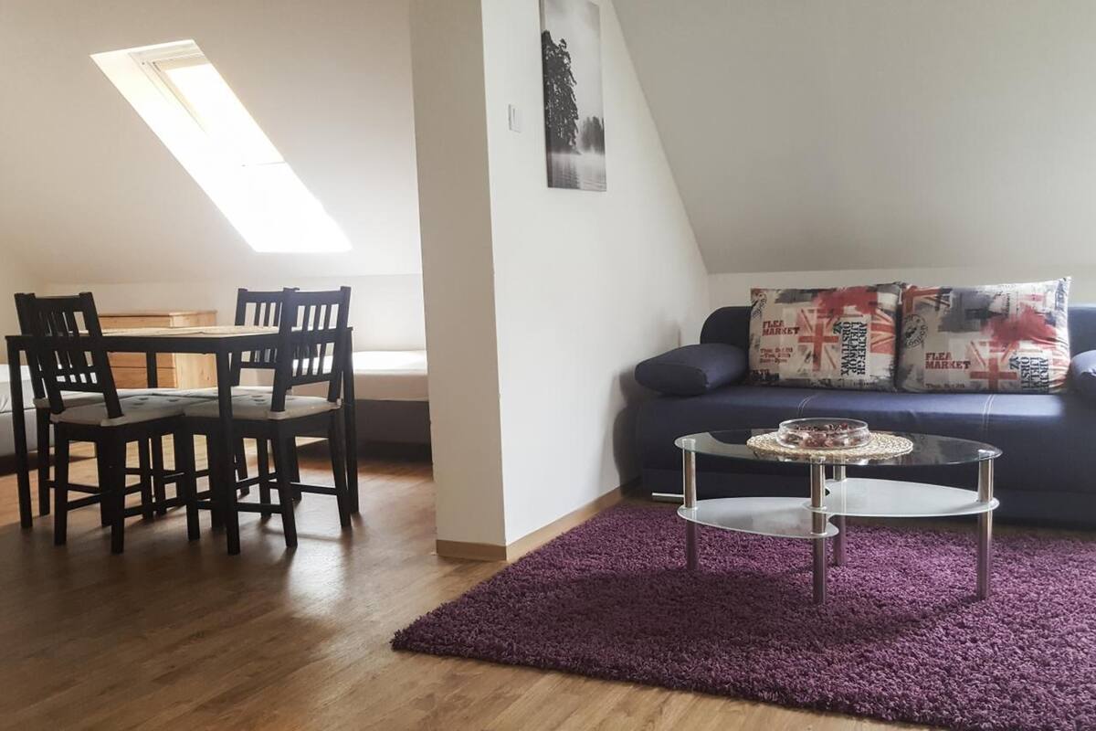 A living room with a dark blue sofa, a small round table, and a purple rug. There is a wooden dining table with chairs and a window with natural light.