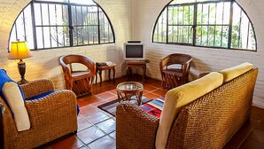 Una sala de estar acogedora con dos ventanas arqueadas, muebles de mimbre, un televisor y una lámpara.