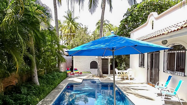 Piscina con sillones y sombrillas, rodeada de palmeras y un edificio blanco con tejado de baldosas.