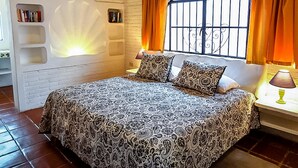 Un dormitorio con una colcha estampada, una ventana con cortinas, una estantería y un estante montado en la pared.