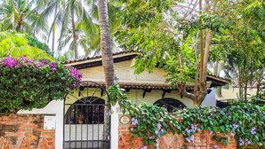 Un edificio de ladrillo con tejado de tejas, entrada con reja y abundante vegetación, incluyendo plantas con flores y palmeras.