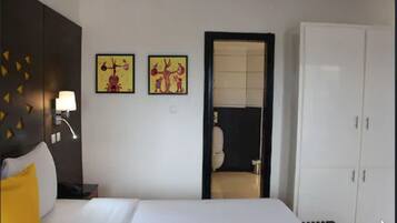 A hotel room with a bed, a nightstand, a lamp, two wall art pieces, a wardrobe, and a bathroom visible through an open door.