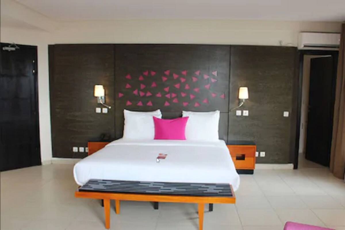 A hotel room with a large bed, two bedside tables, a nightstand, a wall-mounted light fixture, and a decorative wall panel.