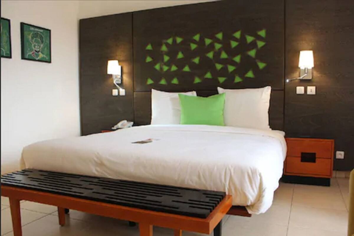 A hotel room with a large bed, two bedside tables, a nightstand, a bench, and a wall with geometric light fixtures.