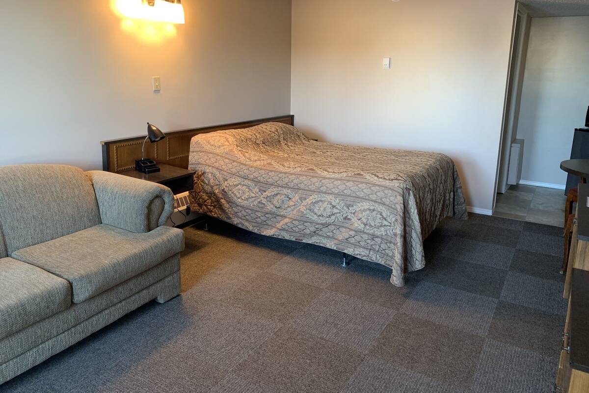 Quarto pequeno e bem arrumado, com cama, sofá, mesa de cabeceira e luminária fixada na parede.