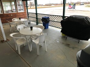 A covered patio area with white plastic chairs and tables, a barbecue grill, and a trash bin. - Airways Motel (Cold Lake)