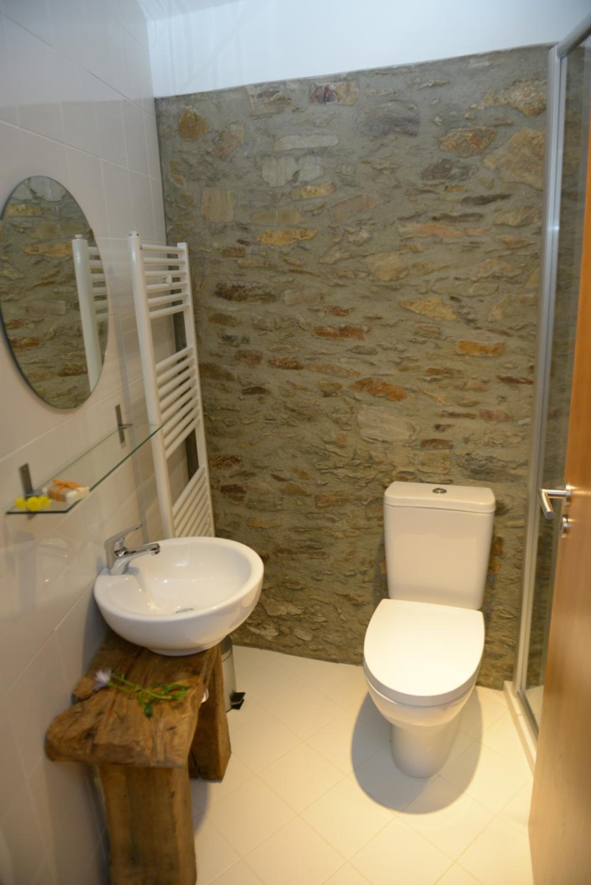 Un baño con pared de piedra, un lavabo blanco, un inodoro y un taburete de madera.
