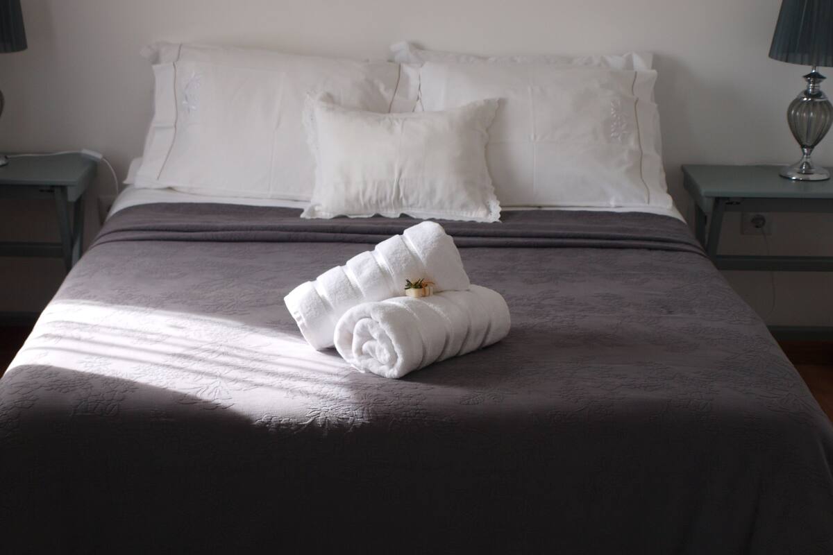 Ein ordentlich bezogenes Bett mit einer grauen Tagesdecke und weißen Kissen, ein gefaltetes Handtuch auf dem Bett sowie eine Lampe auf dem Nachttisch.