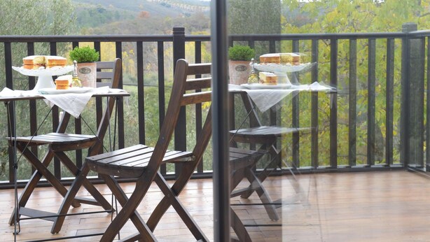 Een balkon met houten stoelen en tafels, plantenbakken met planten, en een mooi uitzicht op heuvels en bomen.