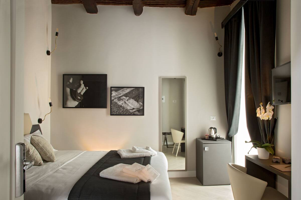 A modern hotel room with a bed, a desk, a chair, a small fridge, and a potted plant.
