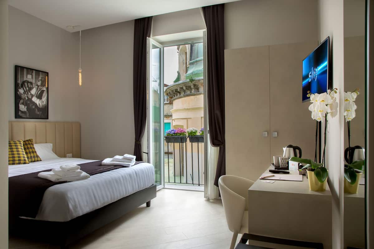 A hotel room with a bed, a desk, a TV, and a balcony with plants.