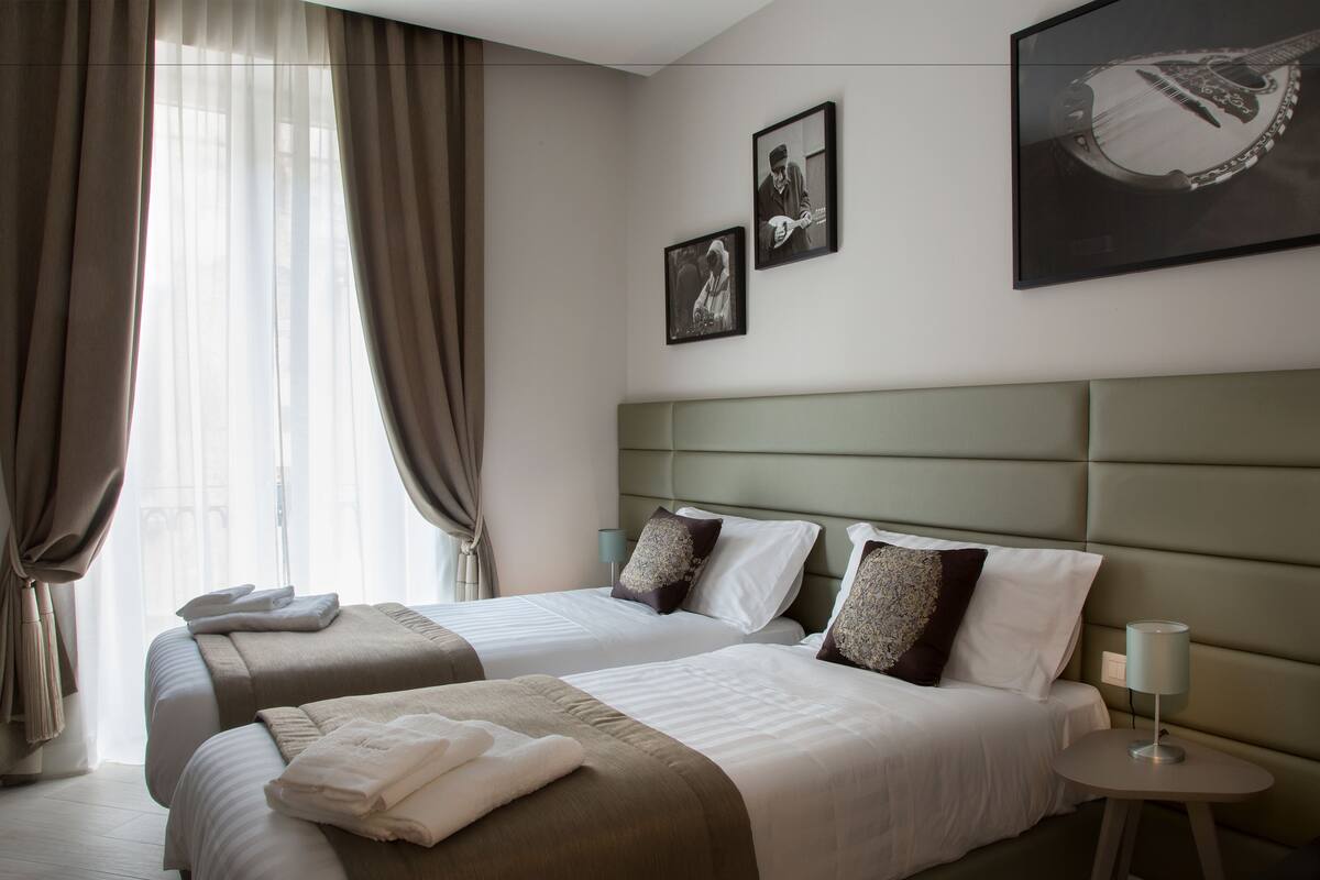 A hotel room with two beds, a headboard, a bedside table, and a window with curtains.