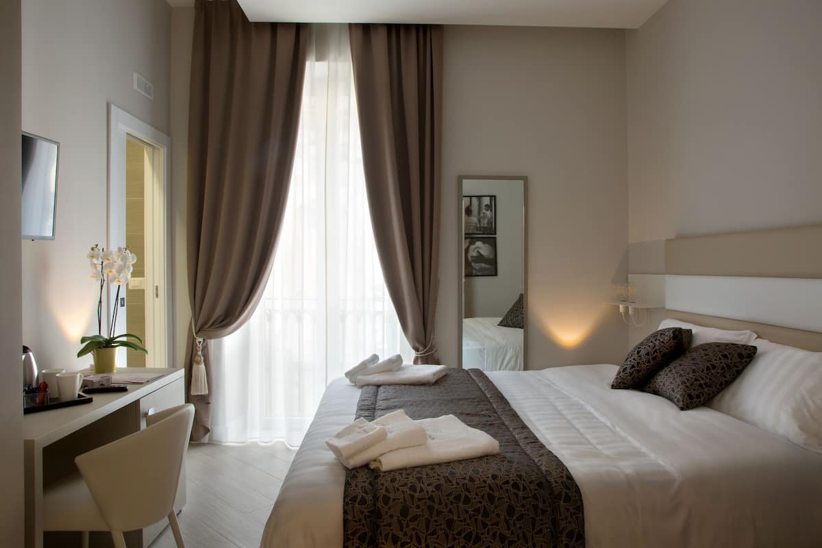 A hotel room with a bed, a desk, a chair, a potted plant, and a window with curtains.