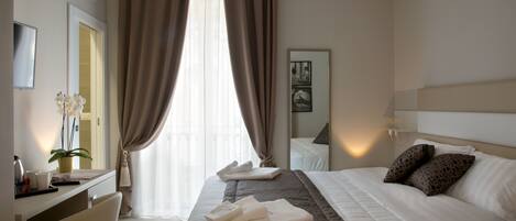 A hotel room with a bed, a desk, a chair, a potted plant, and a window with curtains.