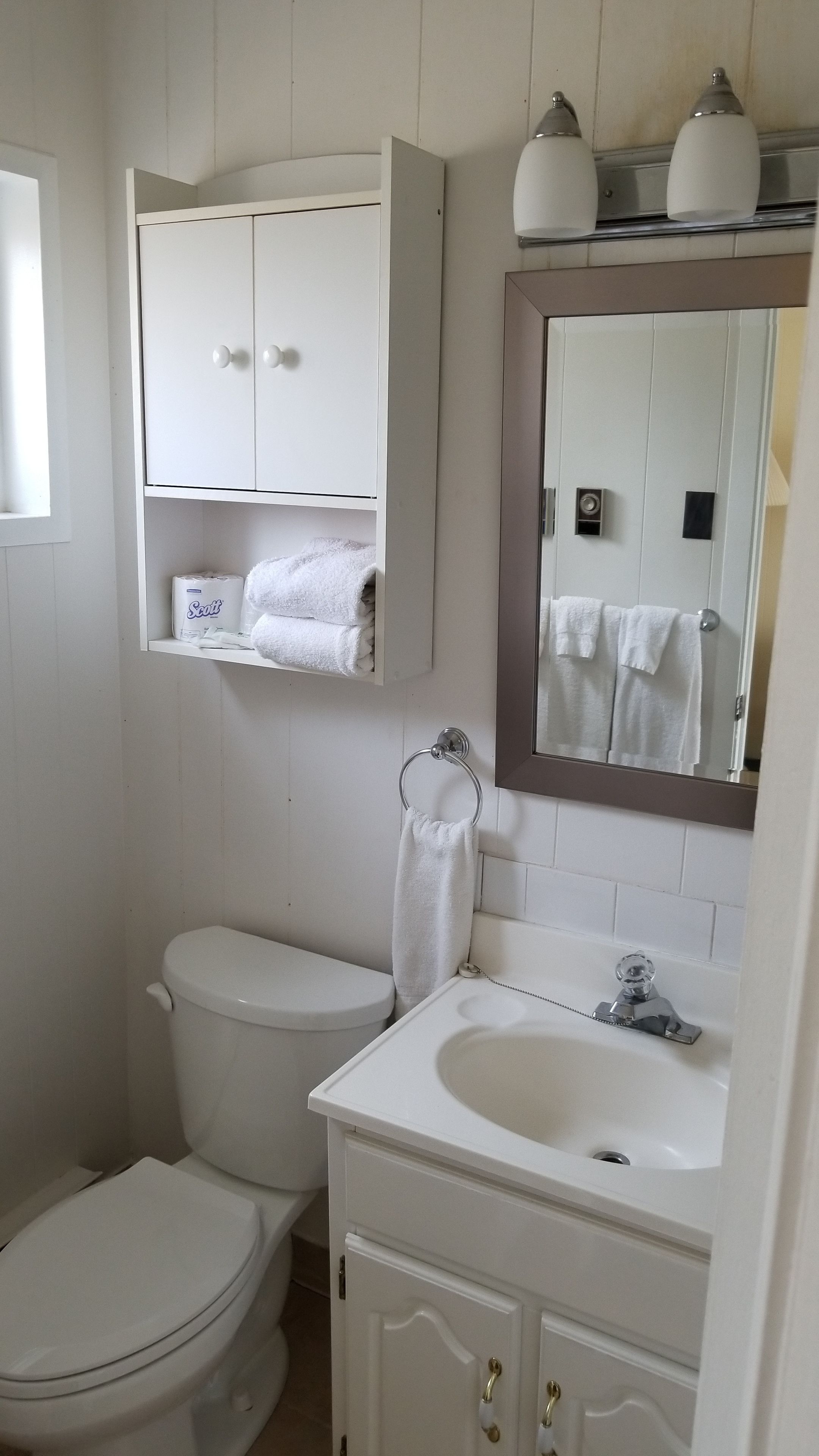 A bathroom with a toilet, sink, and a mirror above the sink. There are towels and a toilet paper roll visible.