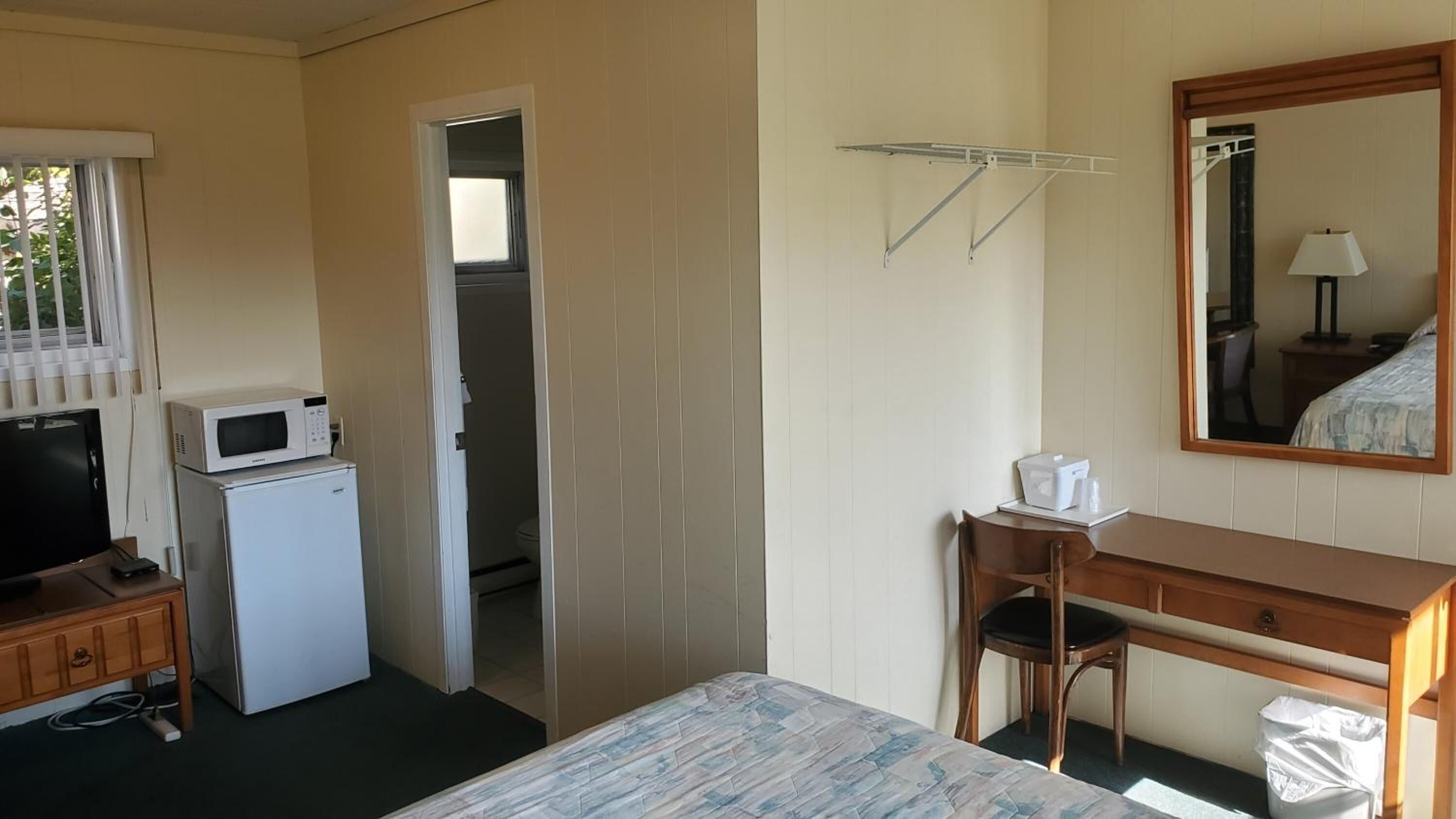 Habitación de hotel compacta con cama, escritorio, silla, microondas, refrigerador pequeño, espejo y ventana con persianas.