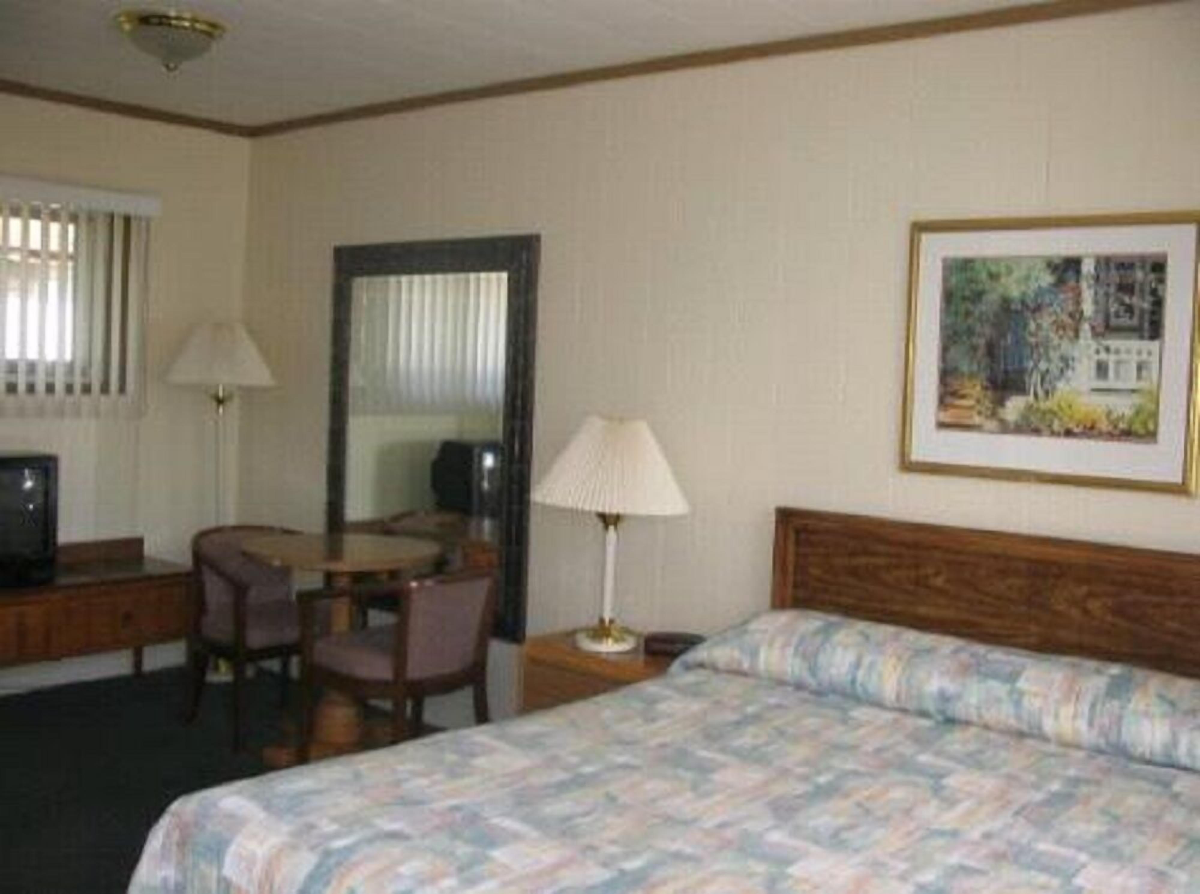 A hotel room with a bed, a desk, a chair, a lamp, a television, and a framed picture on the wall.