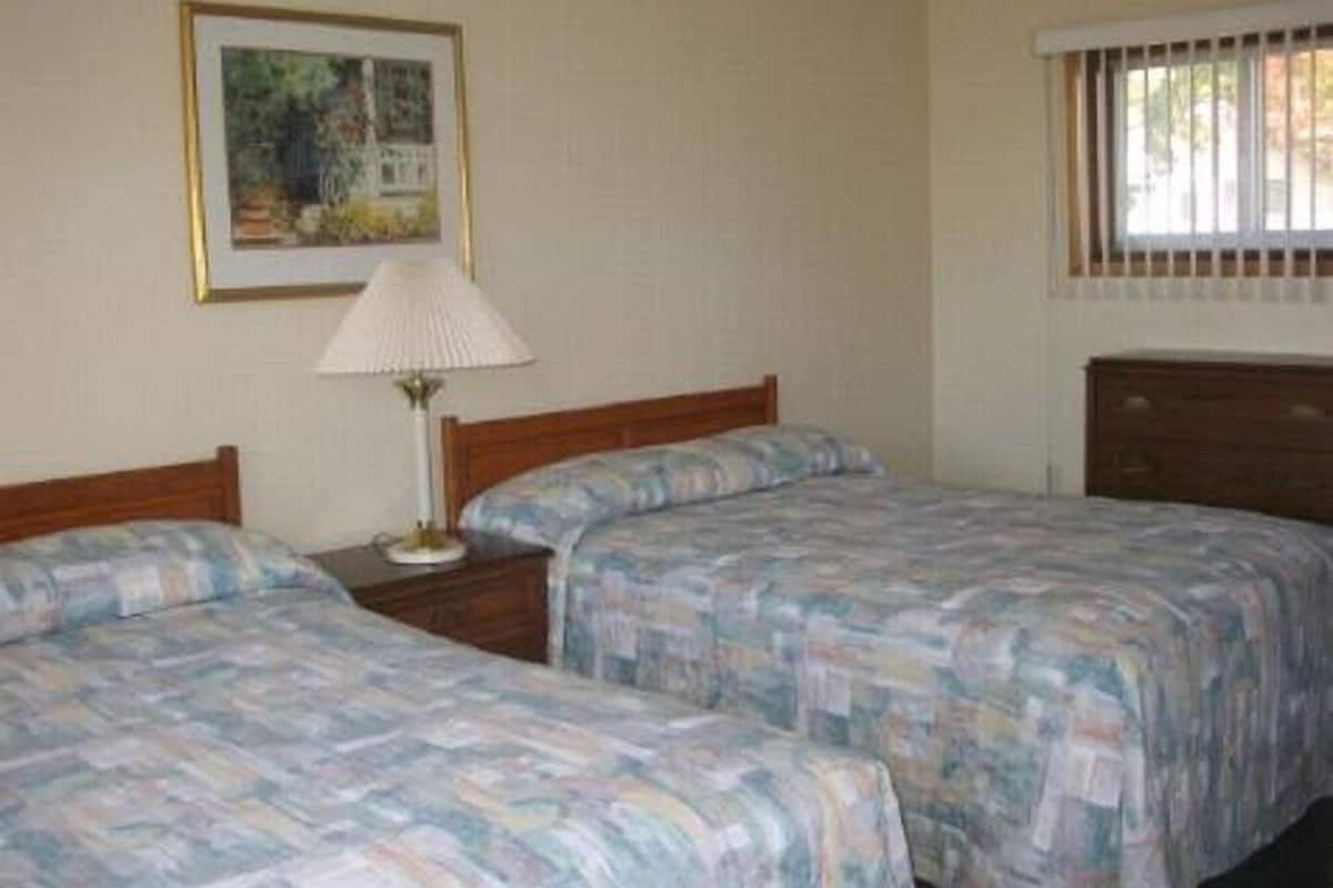 A room with two beds, a bedside table with a lamp, a framed picture on the wall, and a window with bars.