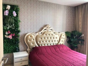 A bedroom with a tufted headboard, a red bedspread, a nightstand, a dresser, and a wall-mounted floral arrangement.