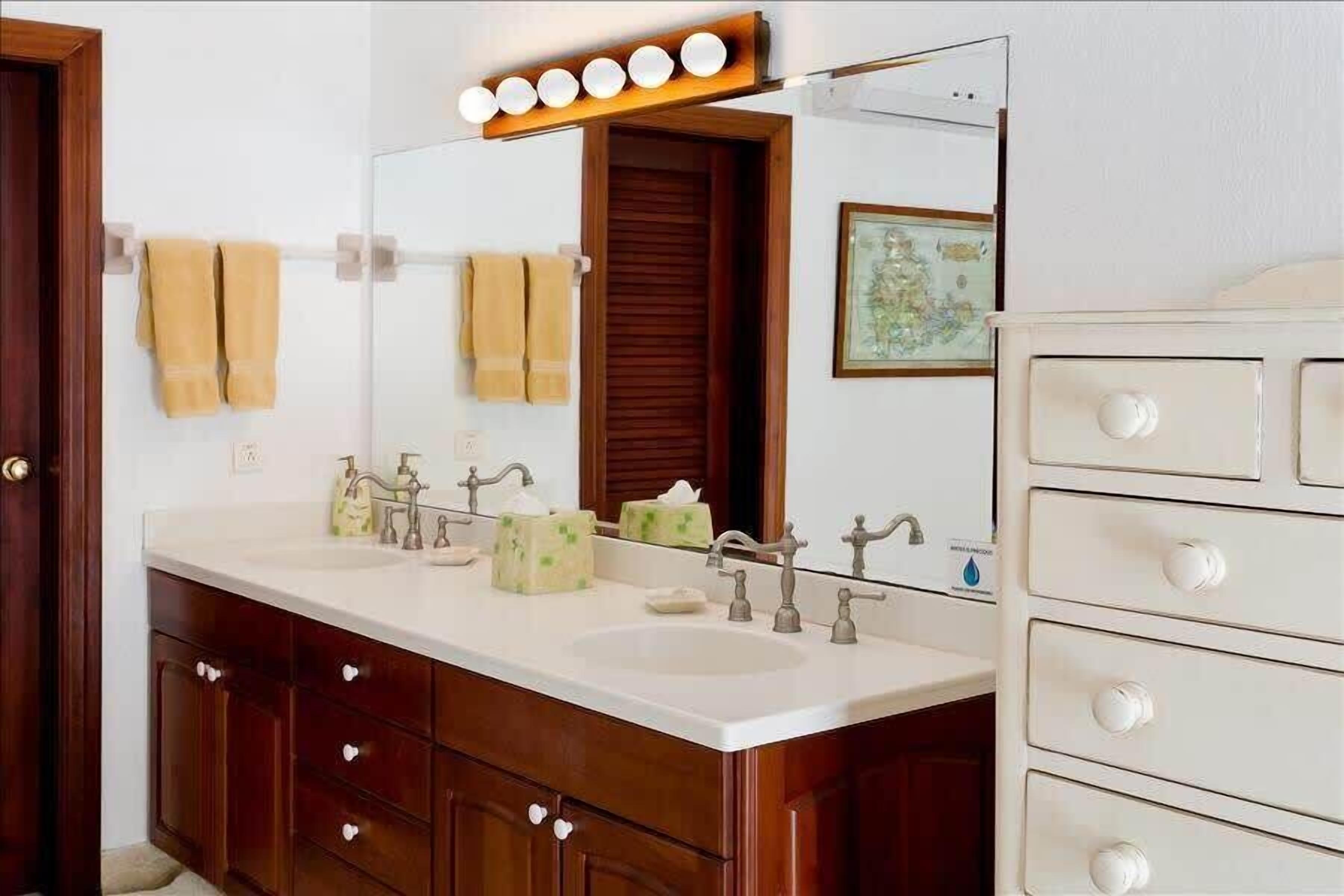 A bathroom with a double sink vanity, wooden cabinets, a mirror with lights, and a framed picture on the wall.