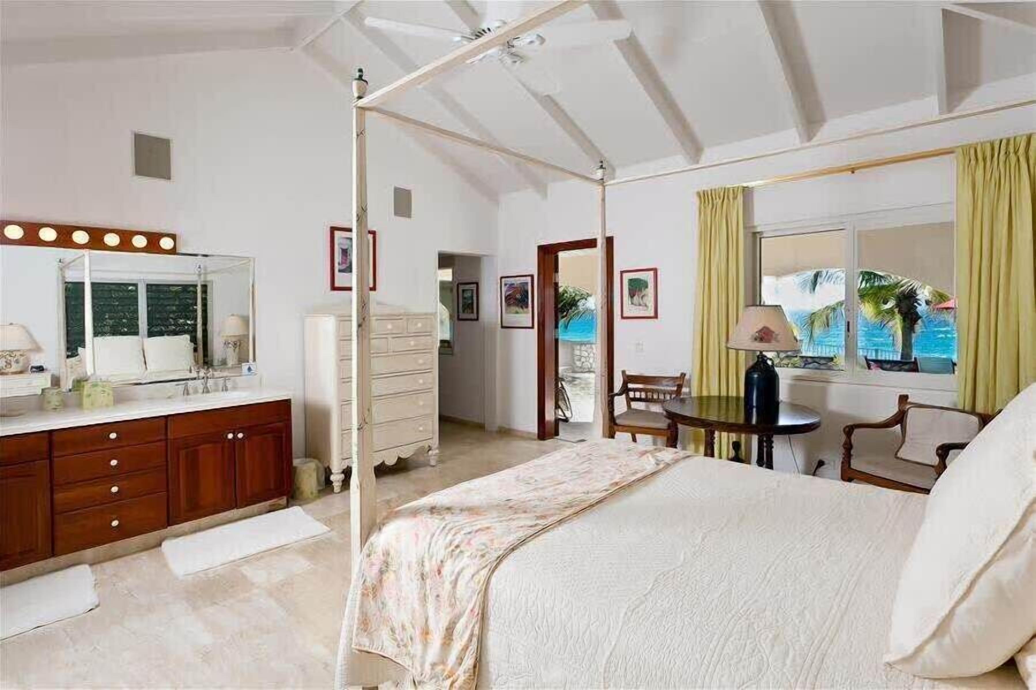 A bedroom with a canopy bed, a vanity with a mirror and lights, a desk with a chair, and a view of the ocean through the window.