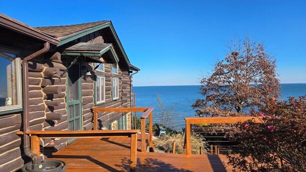 A log cabin with a wooden deck, a metal water spigot, and a view of the sea.