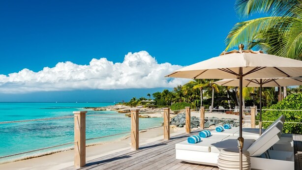 Eine Holzterrasse mit Liegestühlen und Sonnenschirm, mit Blick auf einen türkisblauen Strand und das Meer.