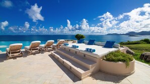 Een zonnig terras met lounge fauteuils en een bank, met uitzicht op een heldere blauwe zee en verre bergen.