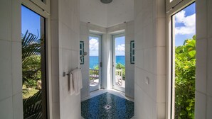 Een smalle betegelde badkamer met een blauwe mozaïekvloer, witte muren en grote ramen met uitzicht op zee.