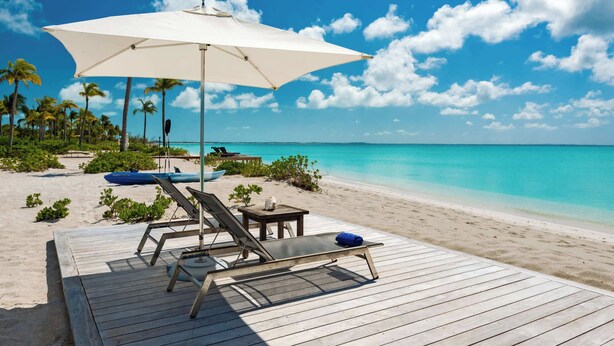 Une terrasse en bois, dotée de chaises longues et d’un grand parasol, qui offre une vue sur une plage aux eaux turquoise et limpides, bordée de palmiers.
